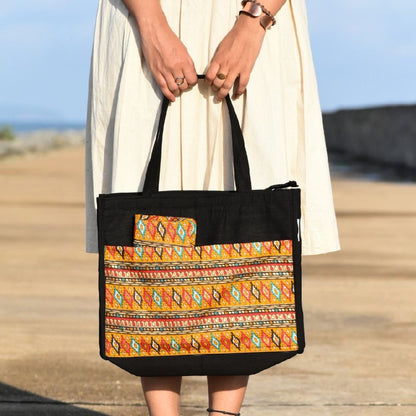 Person holding a black tote bag with colorful pattern on a beach.